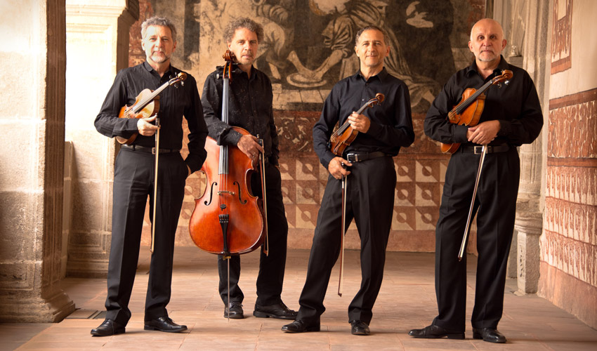 photo of four musicians in black clothes holding their string instruments standing side by side in an old stone hallway lit by natural light