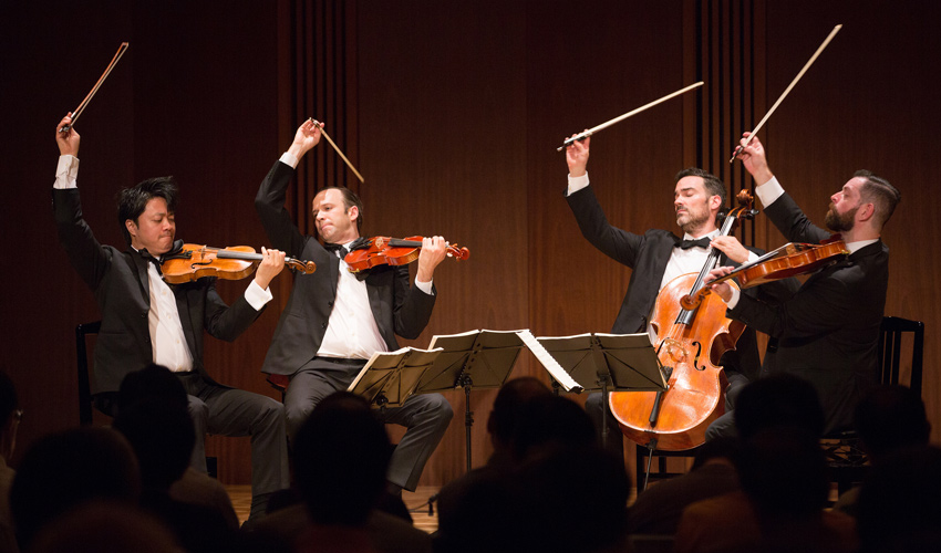 Photo of string quartet musicians wearing tuxedos on stage with their instruments wearing