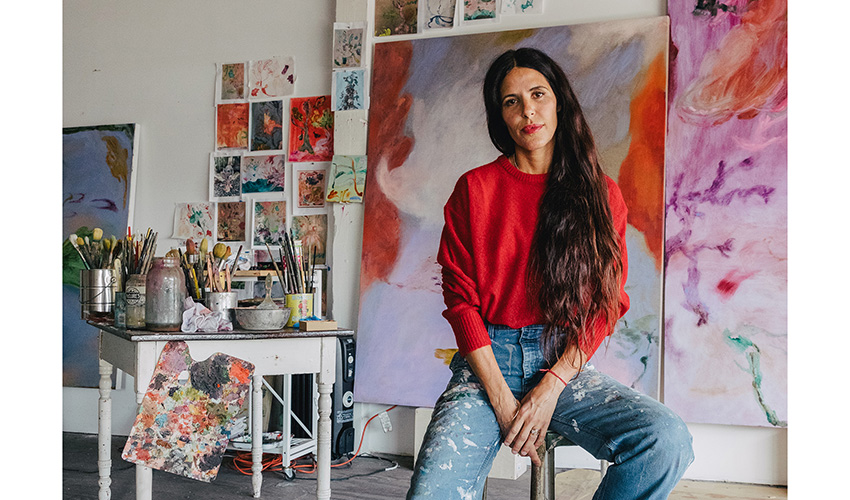 photo of the artist wearing a red sweater and blue jeans, and seated, looking at the camera, in their studio surrounded by their paintings