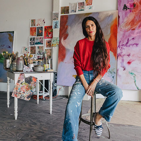 photo of the artist wearing a red sweater and blue jeans, and seated, looking at the camera, in their studio surrounded by their paintings