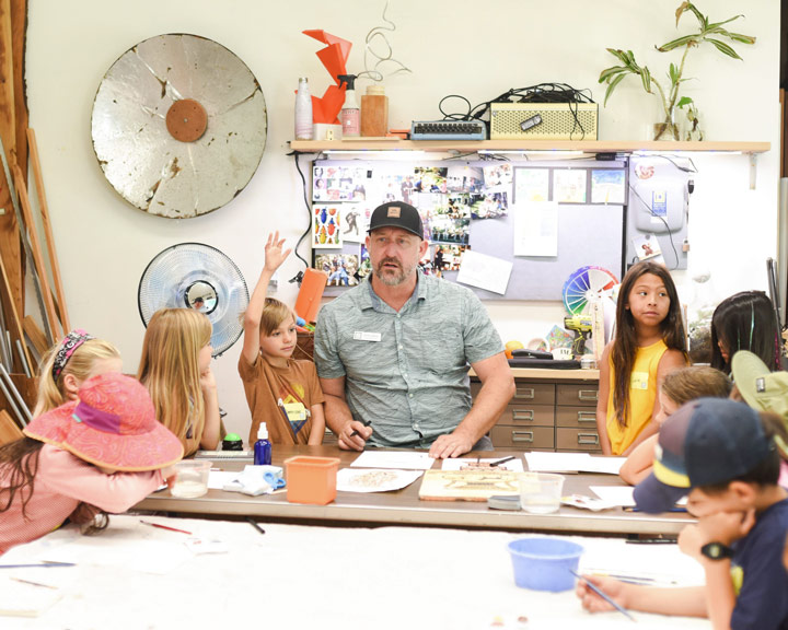 photo of an instructor in baseball cap leading children in arts and crafts projects at a table