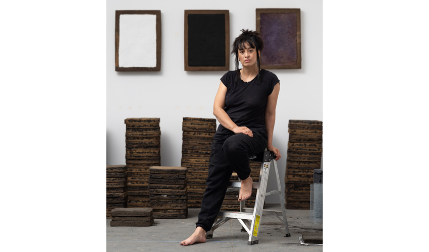 photo of the artist in all black, leaning against a small metal step stool, barefoot, gazing into the camera, against the backdrop of their work