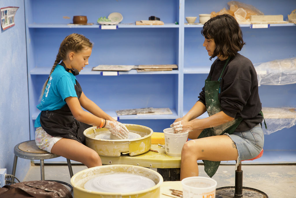 photo of teacher and student in profile sitting at a pottery wheel against a backdrop of a blue bookshelf with crafts on it