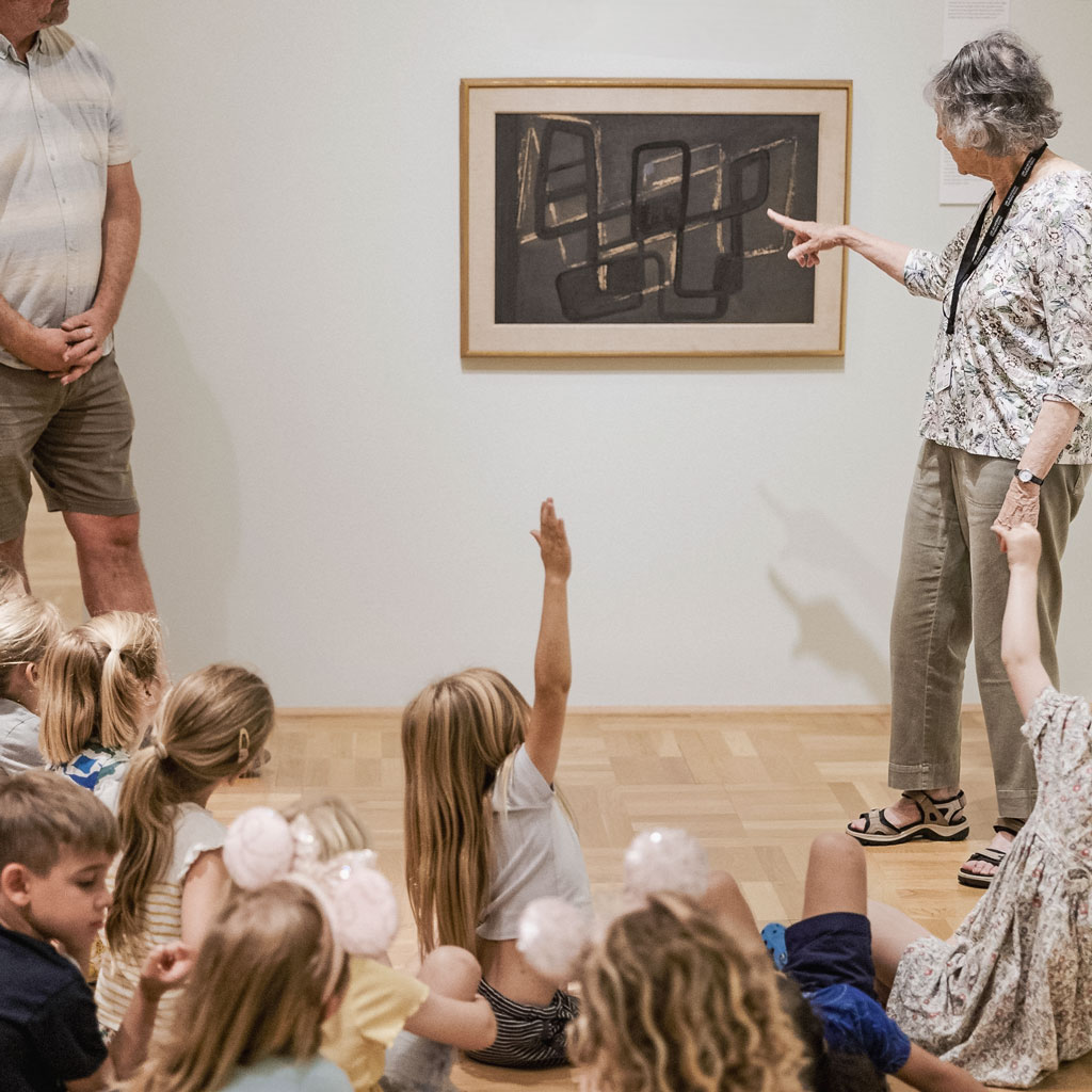 photo of older instructor pointing to a framed work on paper in a museum gallery as seated children look on with one child with long blonde hair raising her hand