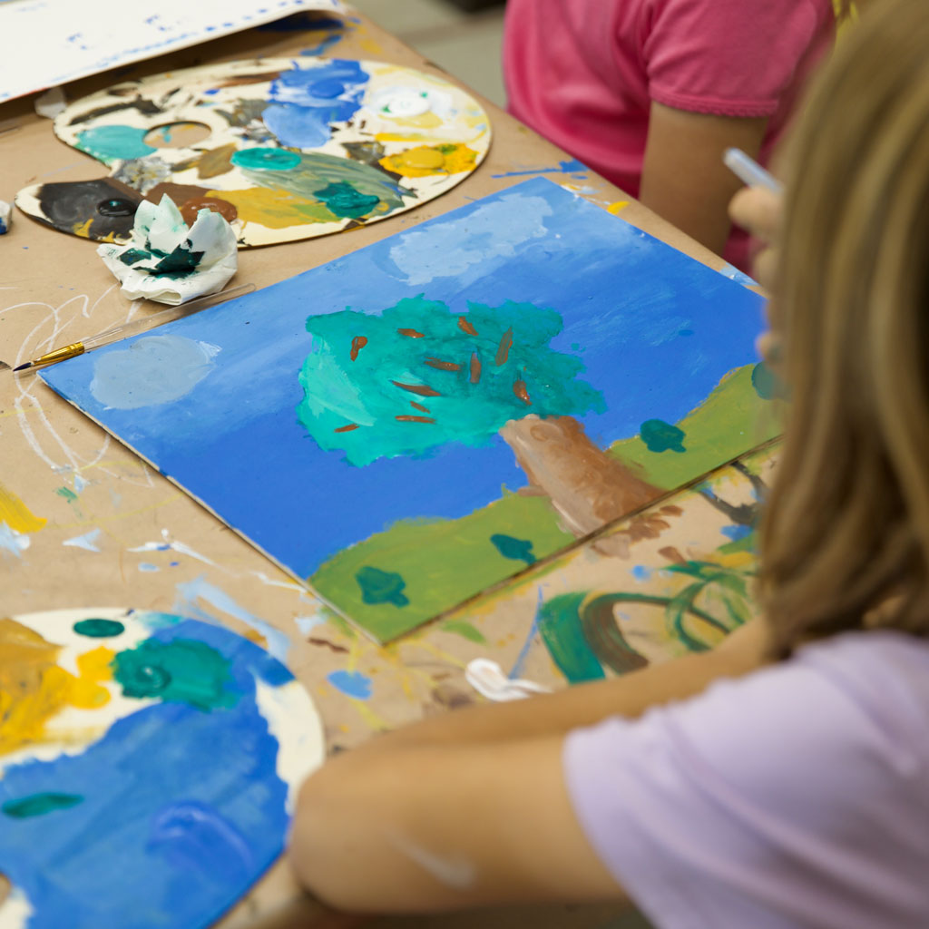 over the shoulder view of a child at a table working on a painting of a tree