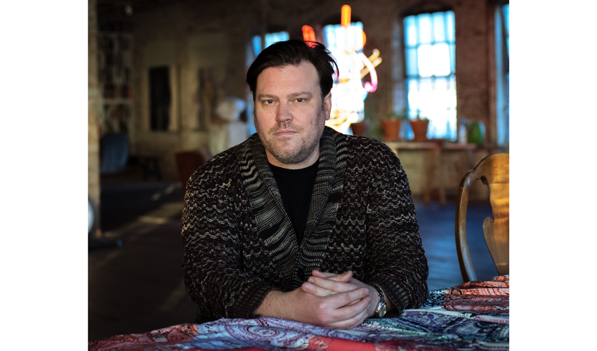 photo of the artist in a dark sweater from the chest up staring into the camera with hands folded in front