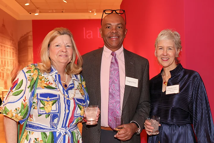 group of three individuals, in cocktail attire at a social event, smiling at the camera