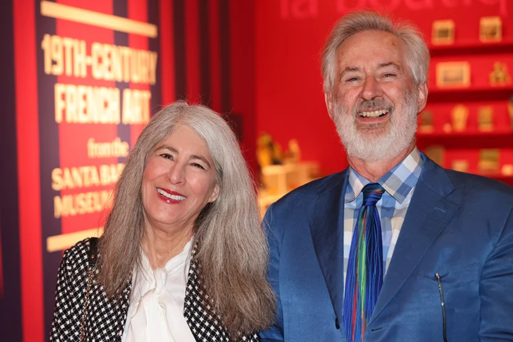 two individuals, in cocktail attire at a social event, smiling at the camera