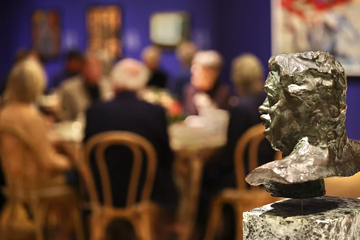 bust of a man in the foreground with a table of people at a society event blurred in the background