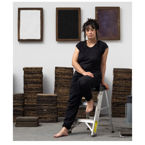 photo of the artist in all black, leaning against a small metal step stool, barefoot, gazing into the camera, against the backdrop of their work