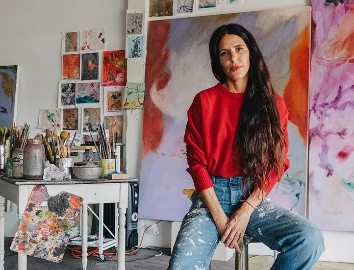 photo of the artist wearing a red sweater and blue jeans, and seated, looking at the camera, in their studio surrounded by their paintings