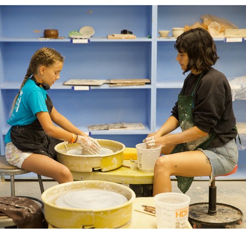 photo of teacher and student in profile sitting at a pottery wheel against a backdrop of a blue bookshelf with crafts on it