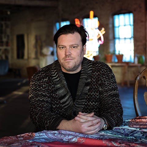 photo of the artist in a dark sweater from the chest up staring into the camera with hands folded in front